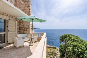 Un balcon aux murs en pierre, un canapé en osier blanc, un parasol vert et une vue sur la mer.