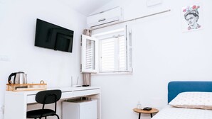 A compact room with a bed, a desk with a chair, a small refrigerator, a television mounted on the wall, and a window with white shutters. - Serious Job Room (Dubrovnik)