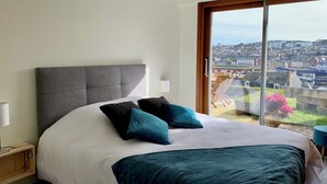 A bedroom with a large bed, a grey headboard, and a view of a cityscape through a large window. - "La Villa du Cap - Sea view - 2/8 travellers (Fécamp)