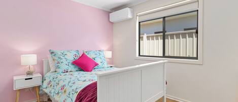 Un dormitorio con una cama, una mesita de noche, una lámpara y una ventana con vista a una cerca blanca.