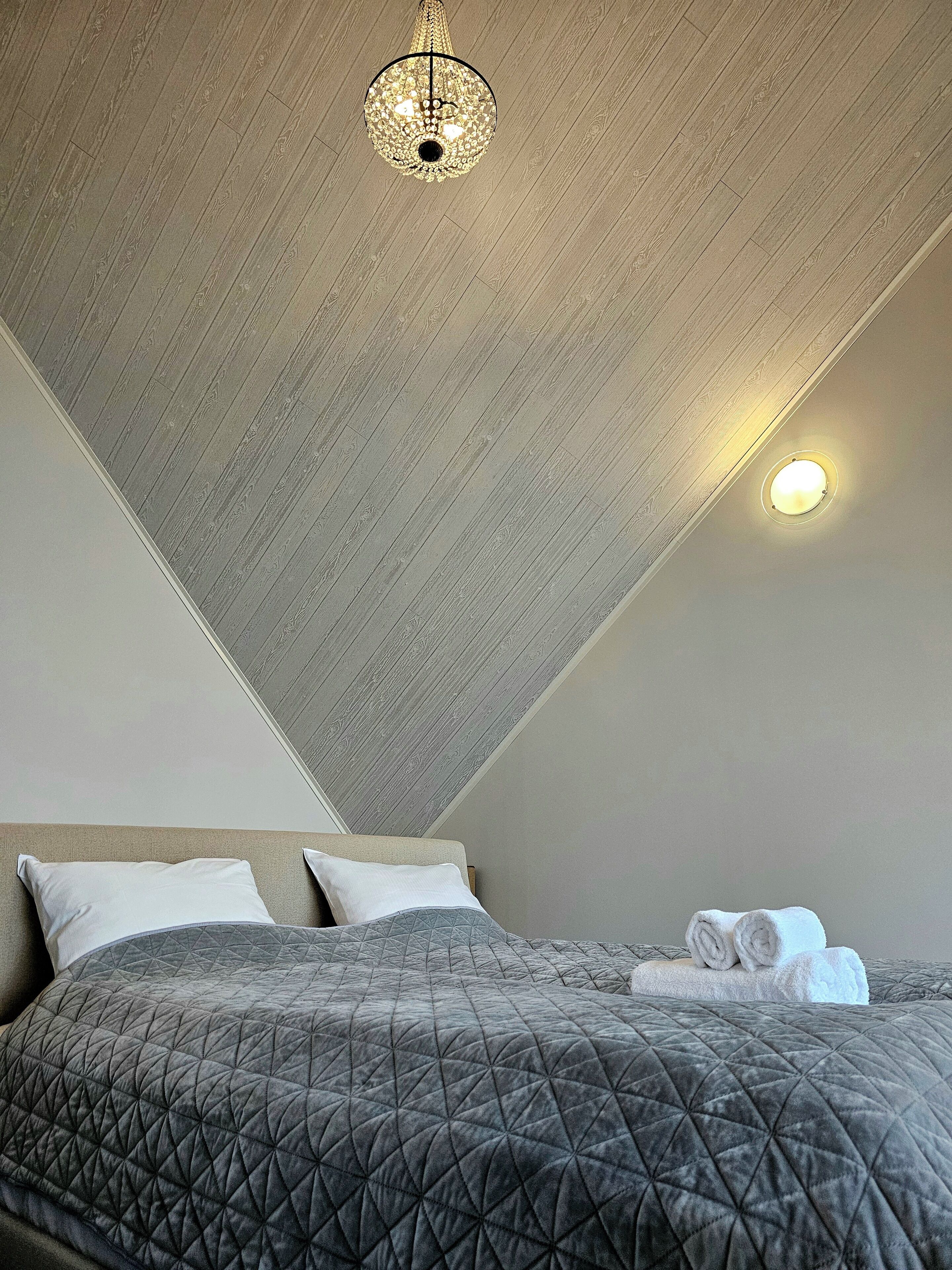 Un dormitorio con techo inclinado, una cama con colcha acolchada gris, dos almohadas y una toalla enrollada sobre la cama.