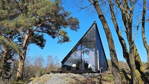 A modern, triangular glass house with a patio area, surrounded by trees and a clear blue sky. - NorGlamp (Hjelmeland)