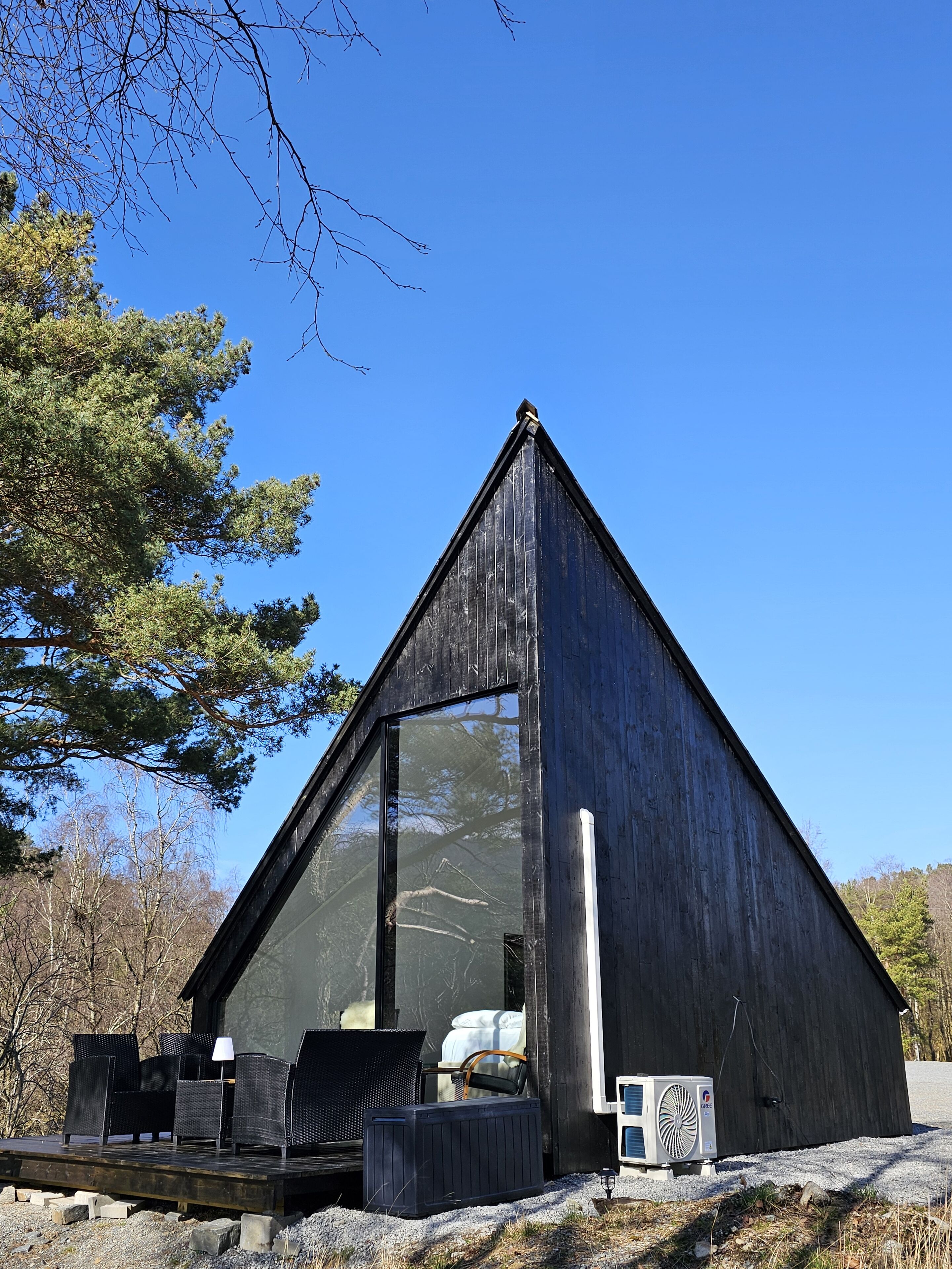 Una cabaña moderna de madera con forma triangular, con un amplio ventanal, amueblada con asientos exteriores y un equipo de aire acondicionado.
