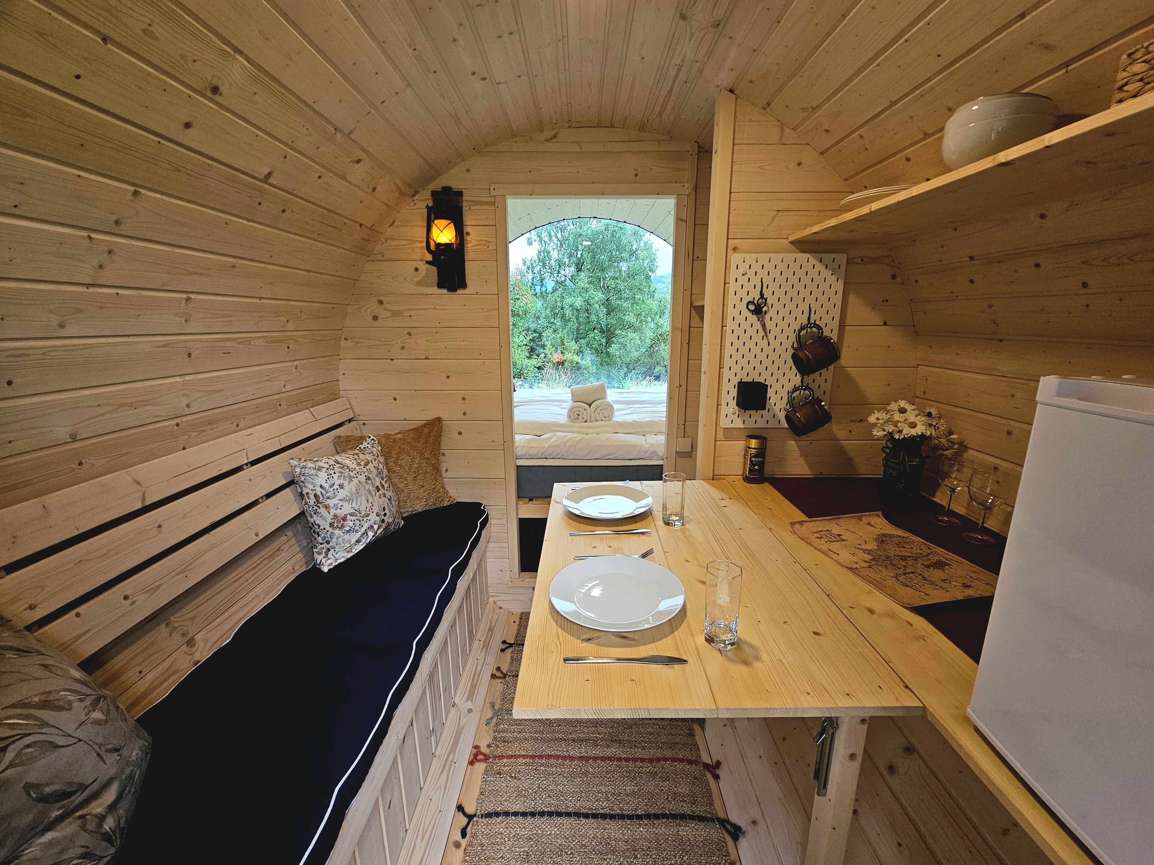Een gezellig houten blokhutinterieur met een bank, een eettafel gedekt met borden, bestek en glazen, en een koelkast.