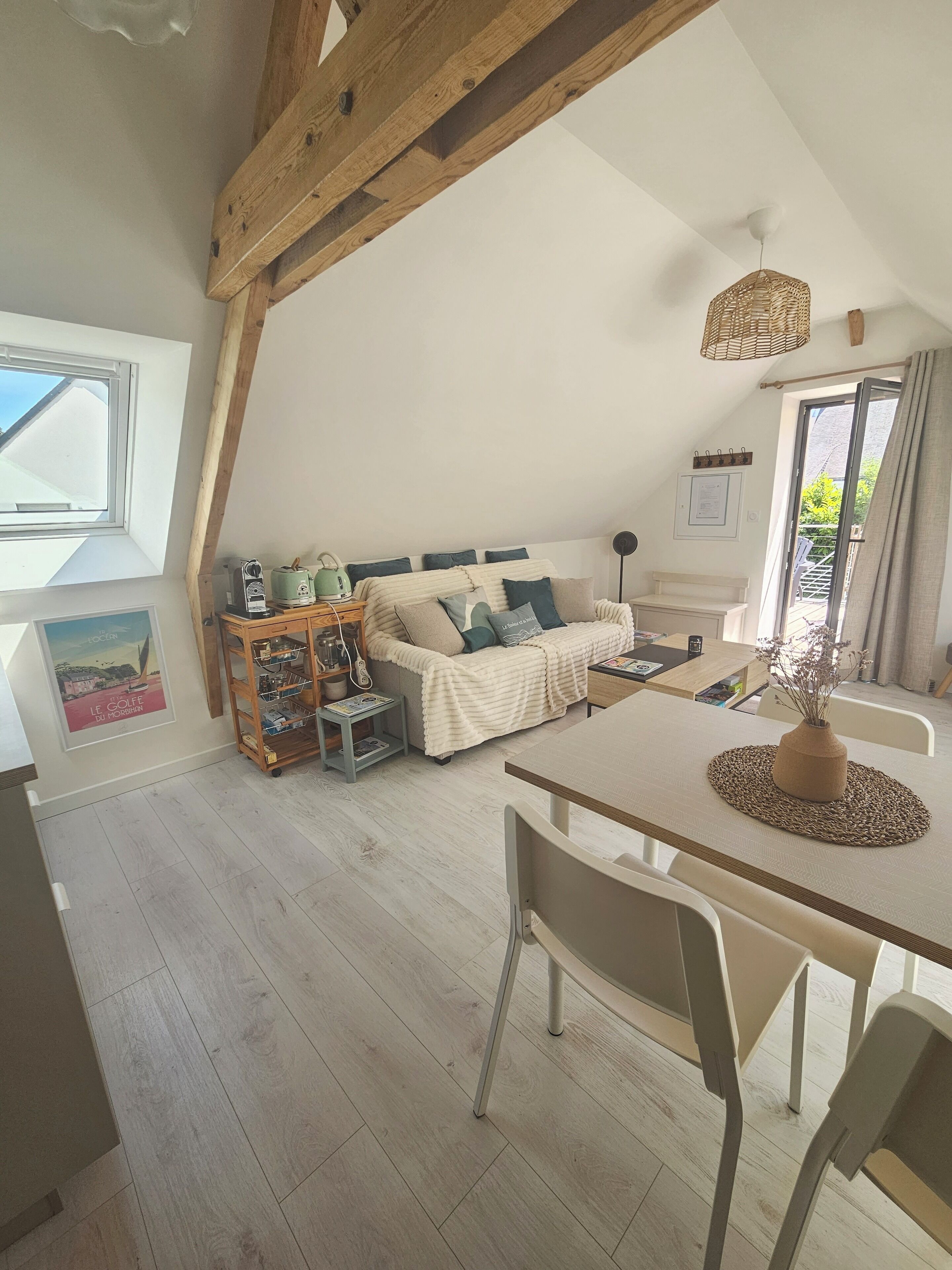 Une chambre à coucher chaleureuse aménagée dans un grenier, avec un plafond en pente, une structure en poutres apparentes, un lit doté d’une tête de lit blanche, une petite table sur laquelle est posé un vase, et une fenêtre donnant sur l’extérieur.