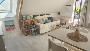 Un dormitorio estilo loft acogedor, con techo inclinado, estructura de vigas de madera, cama con cabecera blanca, una mesita con un florero y ventana con vista al exterior.