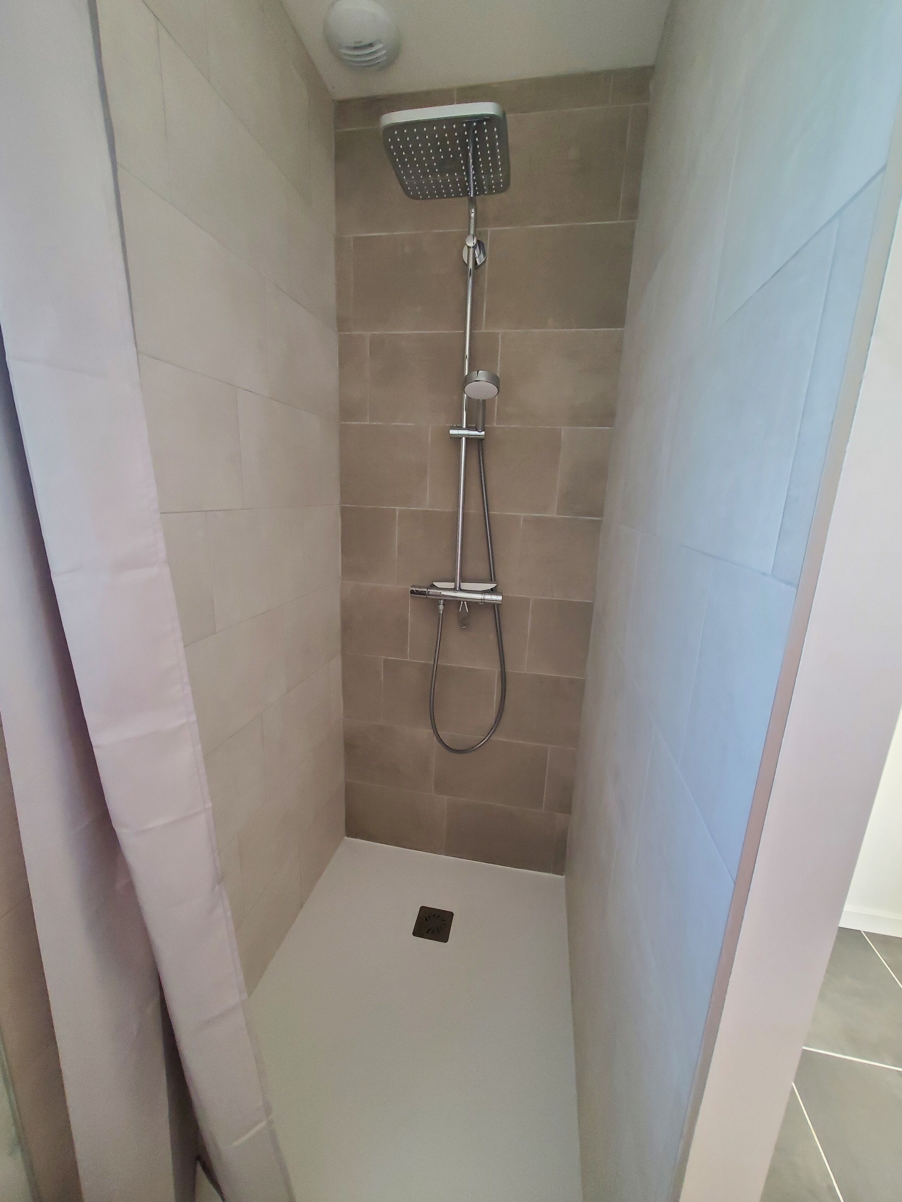 Un petit espace de douche carrelé, équipé d’un pommeau de douche à main et d’un robinet fixé au mur.