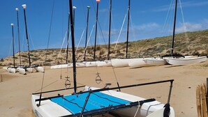 Ein Strand mit einem weißen Boot, das ein blaues Deck und ein Segel hat, umgeben von hohen Masten mit bunten Spitzen, und ein sandiger Strand mit Dünen im Hintergrund.