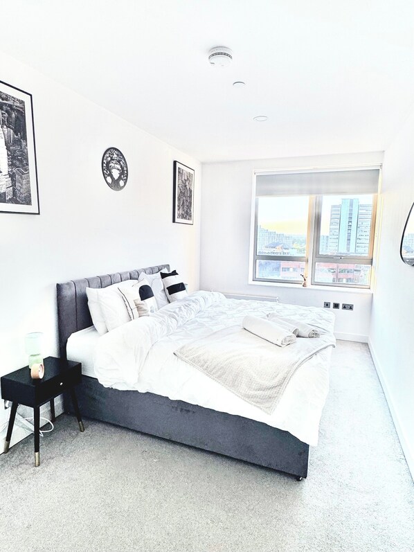 A modern bedroom with a large bed, a nightstand, a window with a city view, and two framed pictures on the wall. - Perfect Stay for Contractors & Families - Sleeps 7 (Bracknell)