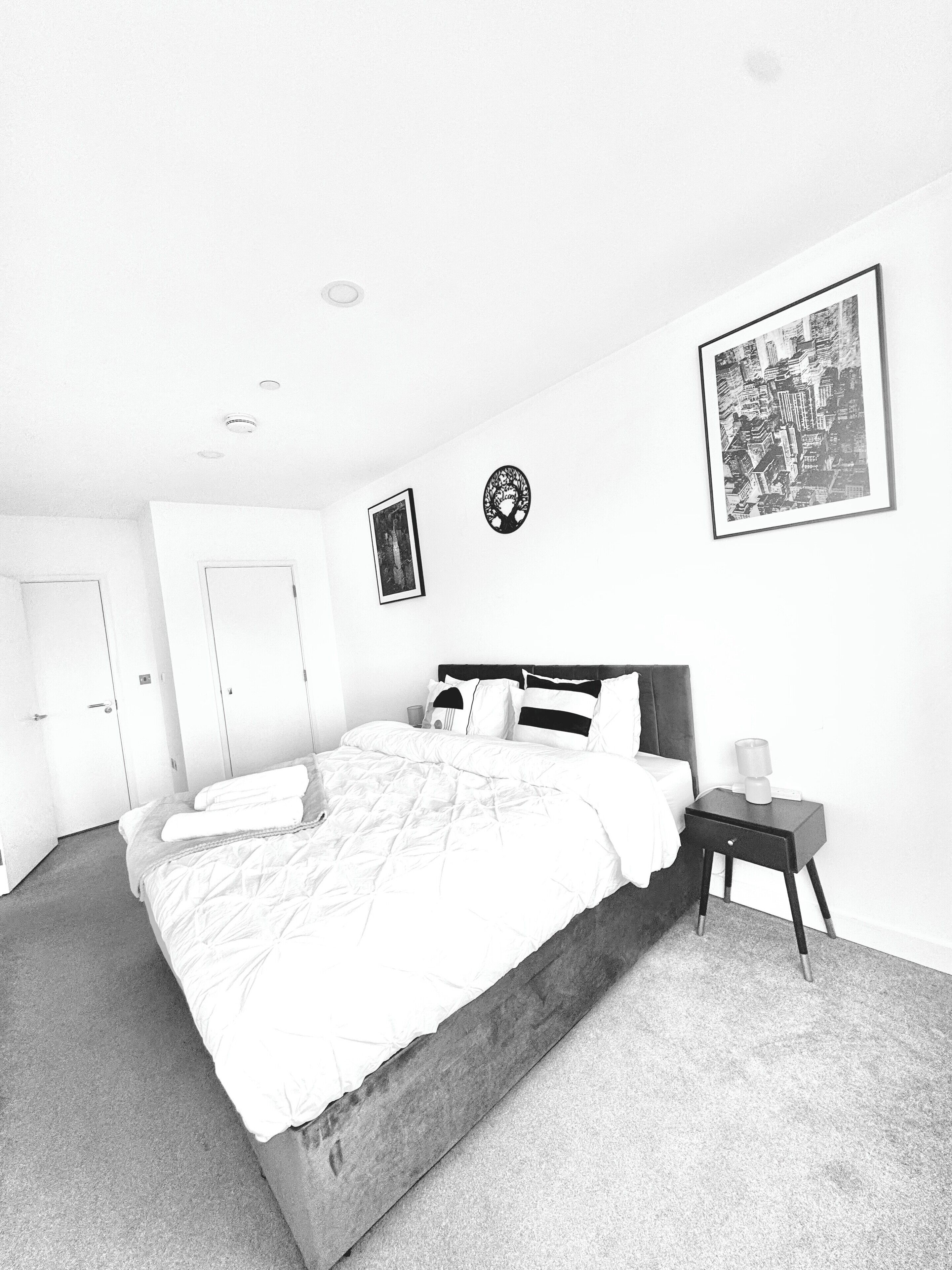 A monochrome hotel room with a large bed, a bedside table, a framed cityscape picture, and a door.