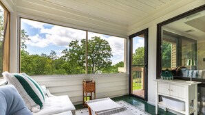 A room with a large window, a white sofa with a green pillow, a small wooden stool, and a white cabinet with drawers.