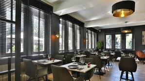 A restaurant with multiple tables and chairs, large windows with blinds, and pendant lighting. - Hôtel L'ermitage (Houffalize)
