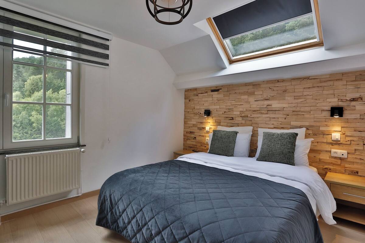 A bedroom with a large bed, a skylight, a wooden headboard, and a view of trees through the window.