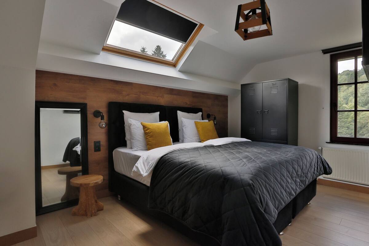 A bedroom with a large bed, a wooden headboard, a skylight, a wardrobe, and a small wooden stool.