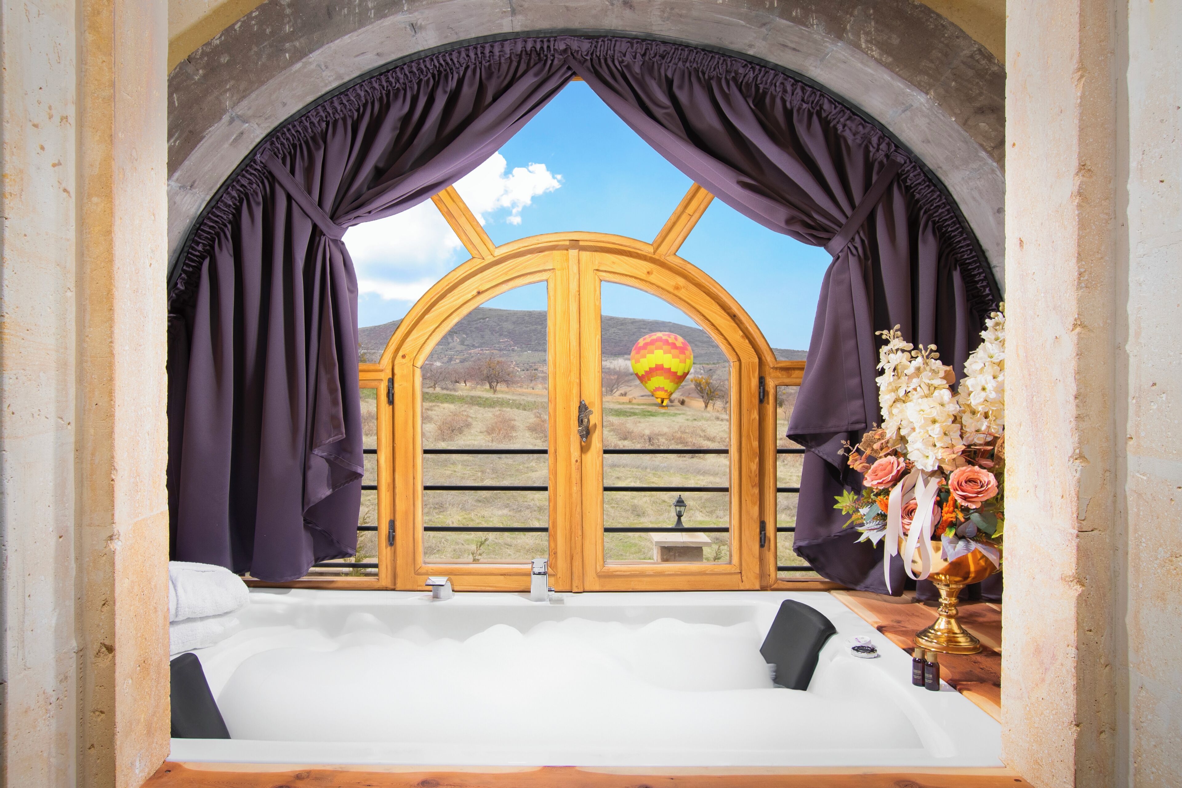 A room with a large arched window, a hot air balloon visible through the window, a bathtub, and a vase with flowers.