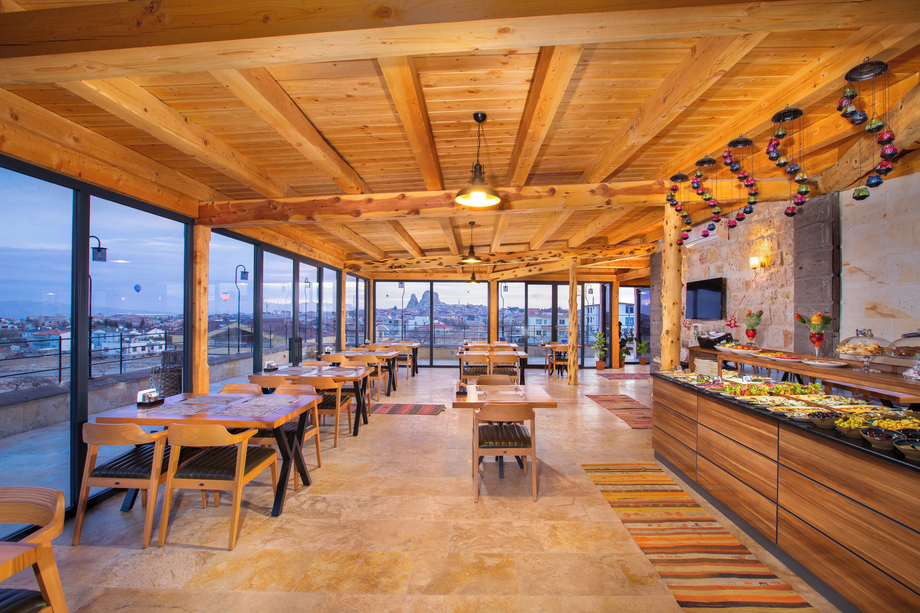 Un amplio comedor con mesas y sillas de madera, un mostrador de buffet con diversos alimentos, y grandes ventanales que ofrecen vistas de la ciudad.