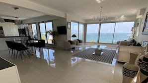A spacious living area with a large window overlooking the sea, a dining area with modern furniture, and a kitchen with a view.