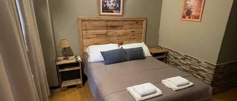 A bedroom with a wooden headboard, a bed with two pillows, a nightstand with a lamp, and two framed pictures on the wall.