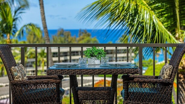 A balcony with wicker chairs and a glass table, overlooking a beach with palm trees and the ocean.