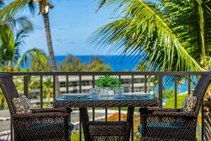 A balcony with wicker chairs and a glass table, overlooking a beach with palm trees and the ocean. - Kihei Akahi D-510 1 Bedroom Condo (Kihei)