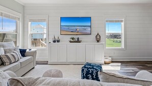 A spacious living room with a large TV mounted on the wall, a comfortable sofa, and a view of the beach through the windows.