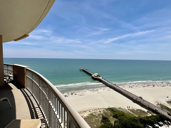 Ein Strand mit einem Steg, der sich ins Meer erstreckt, feiner Sand und ein Balkon mit Blick auf die Szene.