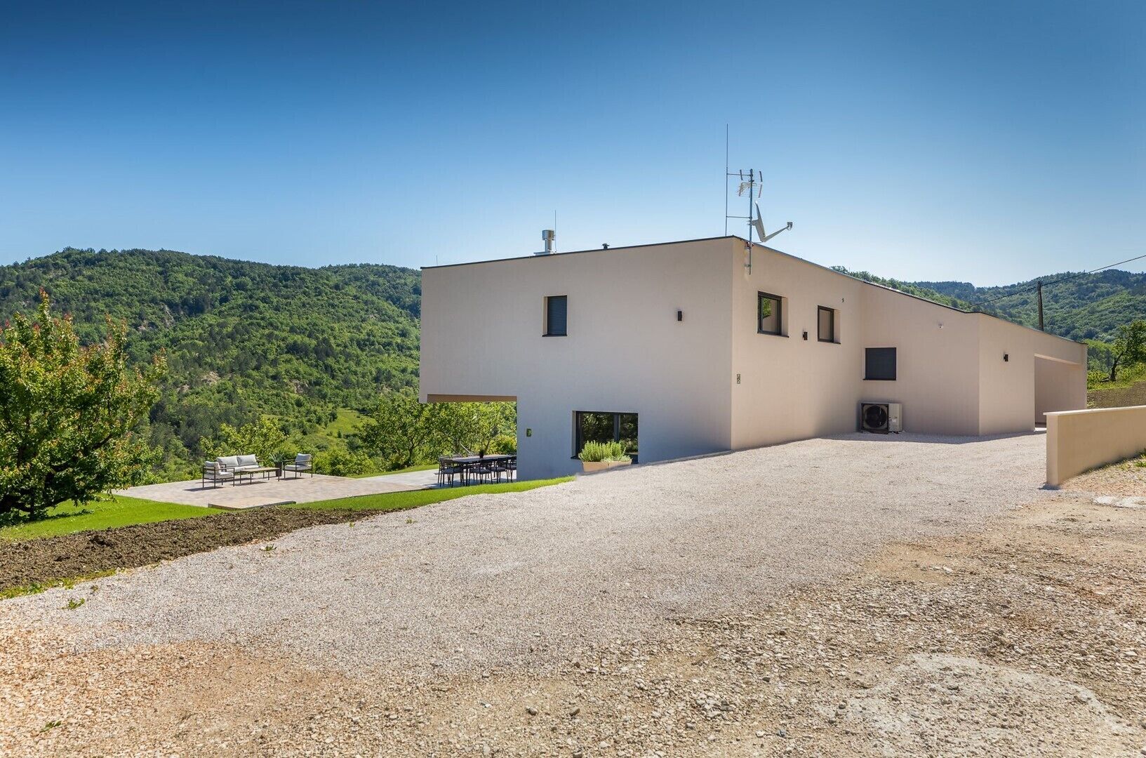 A modern two-story building with a flat roof, a gravel driveway, and a small patio area with outdoor furniture.
