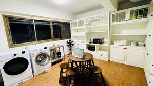 Cocina compacta con lavadora, ventilador, microondas y una mesa con taburetes.