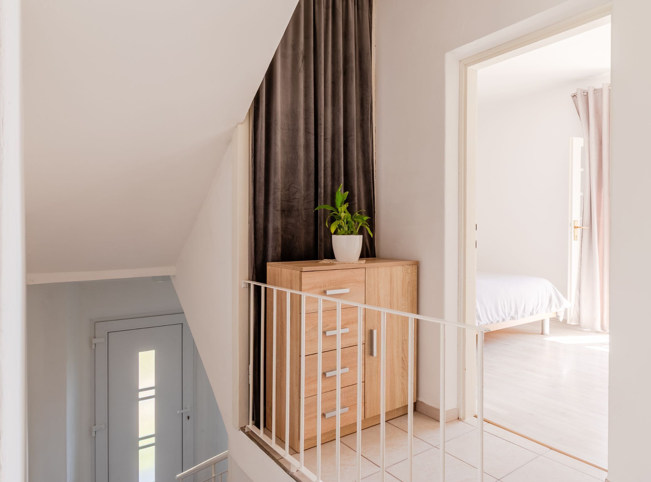 A bedroom with a bed, a wooden cabinet, a potted plant, and a view of another room.