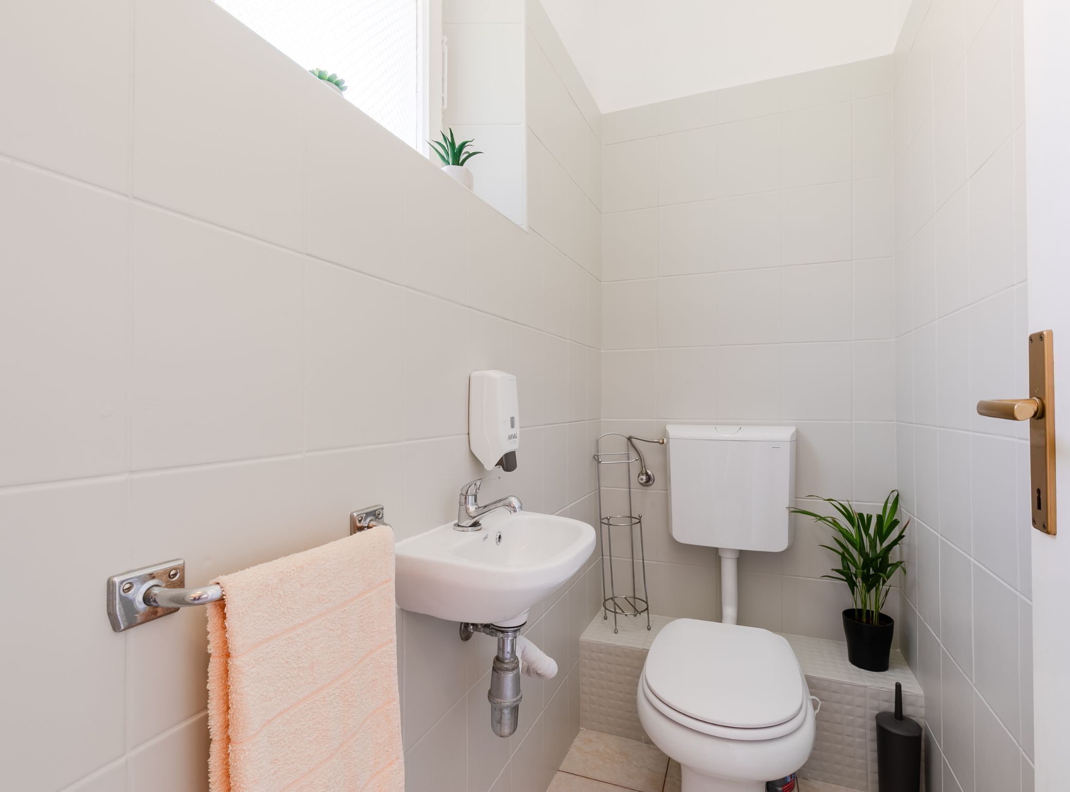 Ein kleines, modernes Badezimmer mit weißen Fliesen, einem Waschbecken, einer Toilette, einem Handtuchhalter, einer Seifenspender und einer Topfpflanze.