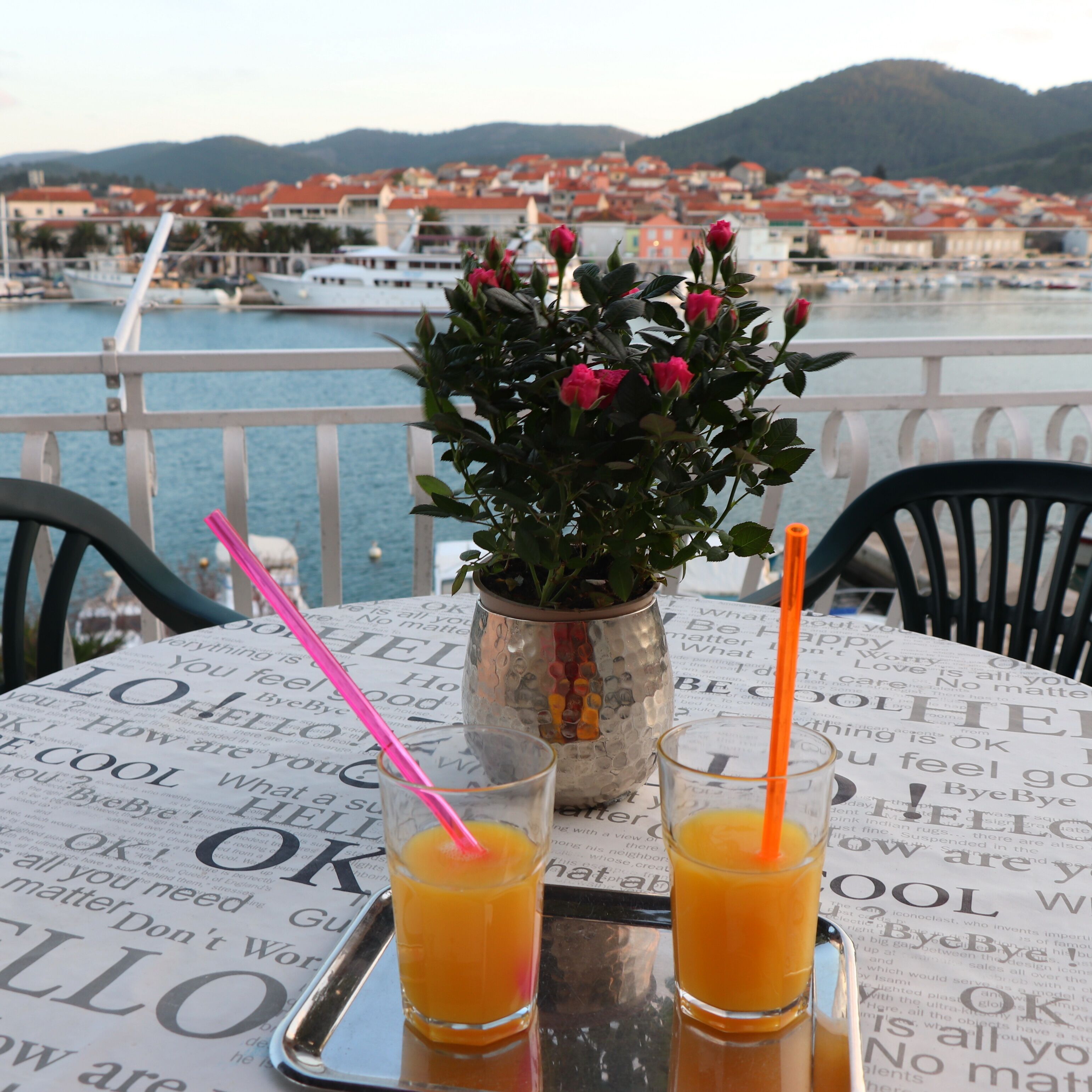A table with two glasses of orange juice, a vase with pink flowers, and a view of a harbor with boats and buildings in the background.