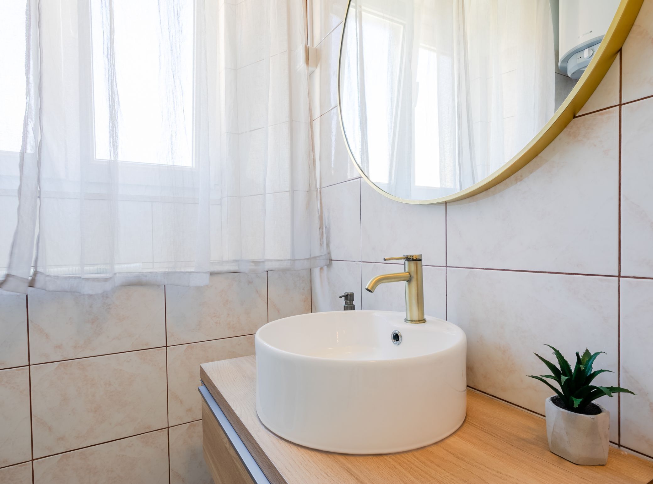 A bathroom with a round white sink, a gold faucet, a mirror, and a potted plant.