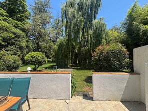 Een terras met een tafel en stoelen, omringd door groen en een grote, hangende wilg.
