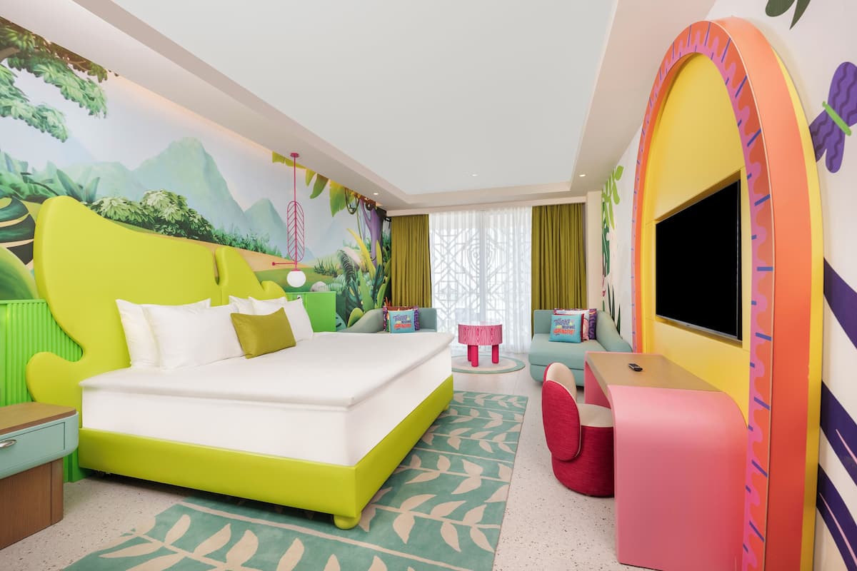 Habitación de hotel luminosa y colorida con una cama grande, televisor, escritorio y un mural colorido en la pared.