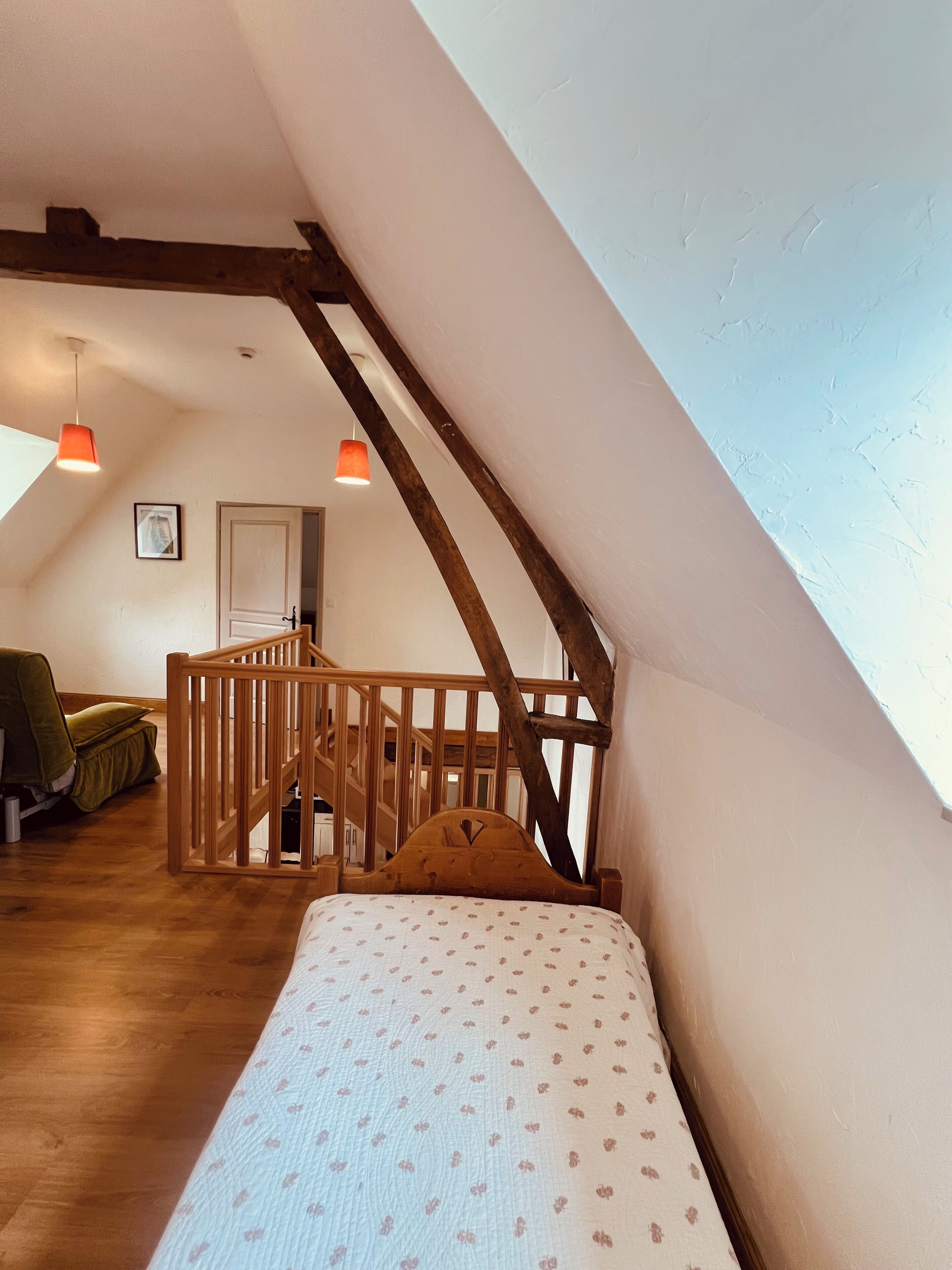 A bedroom with a sloped ceiling, wooden beams, a bed with a patterned bedspread, a chair, and a staircase leading to an upper level.