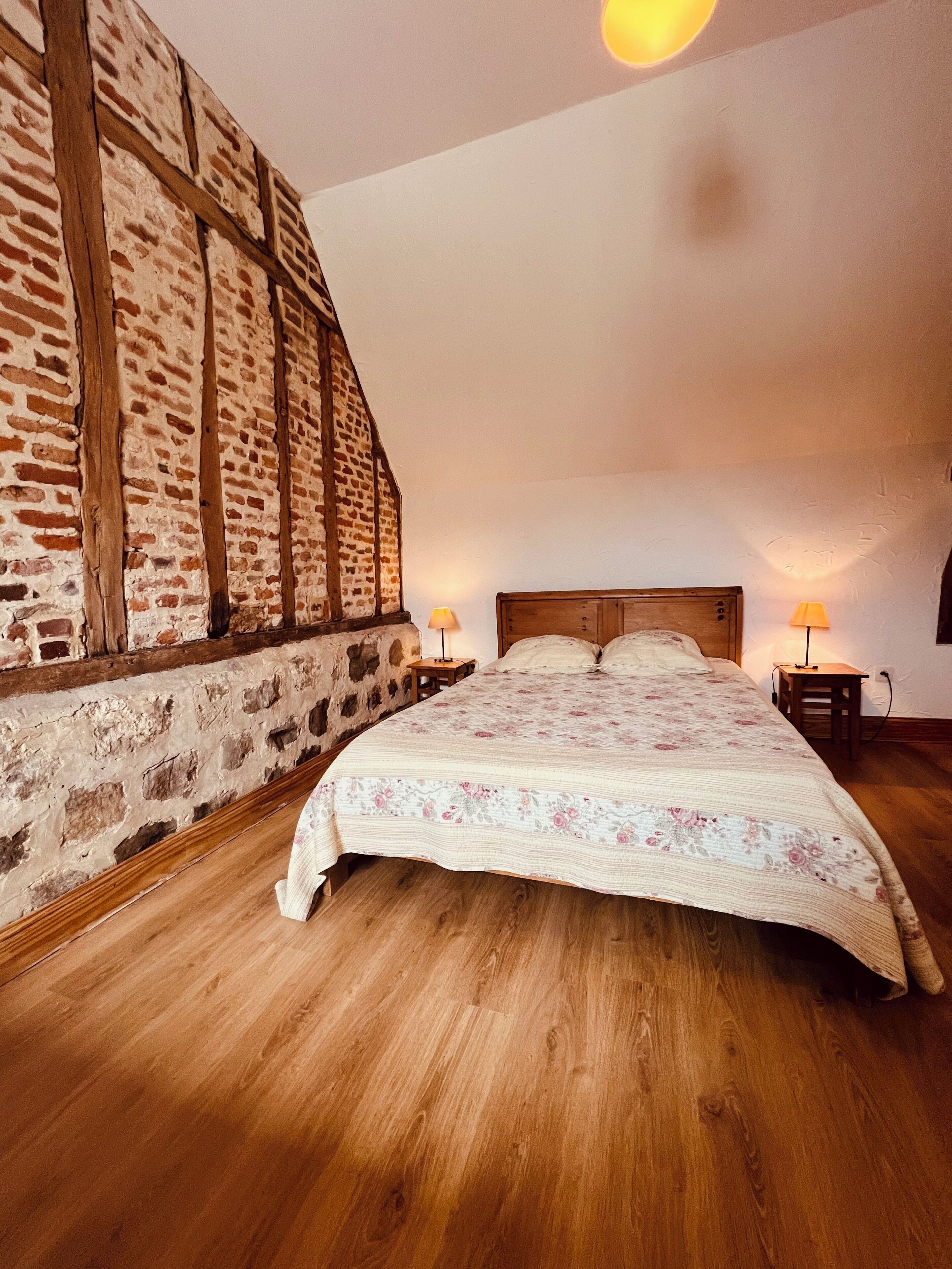 A bedroom with a wooden floor, a bed with a floral bedspread, two bedside lamps, and a brick wall with wooden beams.
