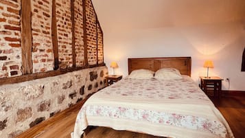 A bedroom with a wooden floor, a bed with a floral bedspread, two bedside lamps, and a brick wall with wooden beams.