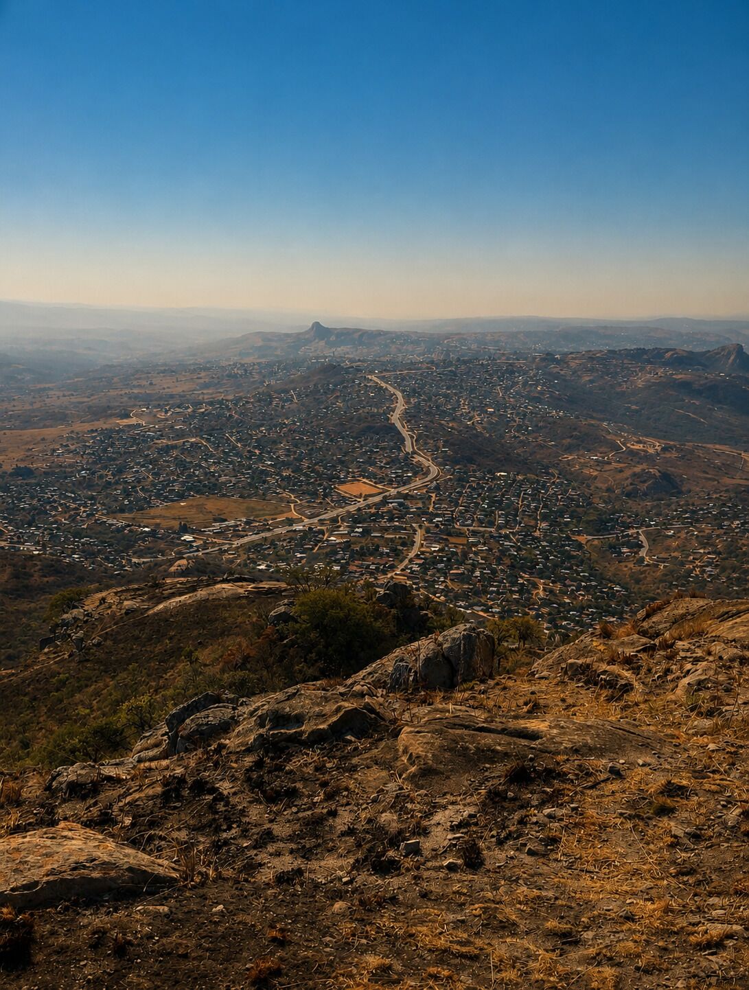 Vista panorámica de una ciudad desde un lugar elevado, con una carretera serpenteante en el centro, rodeada de terreno rocoso y vegetación escasa.
