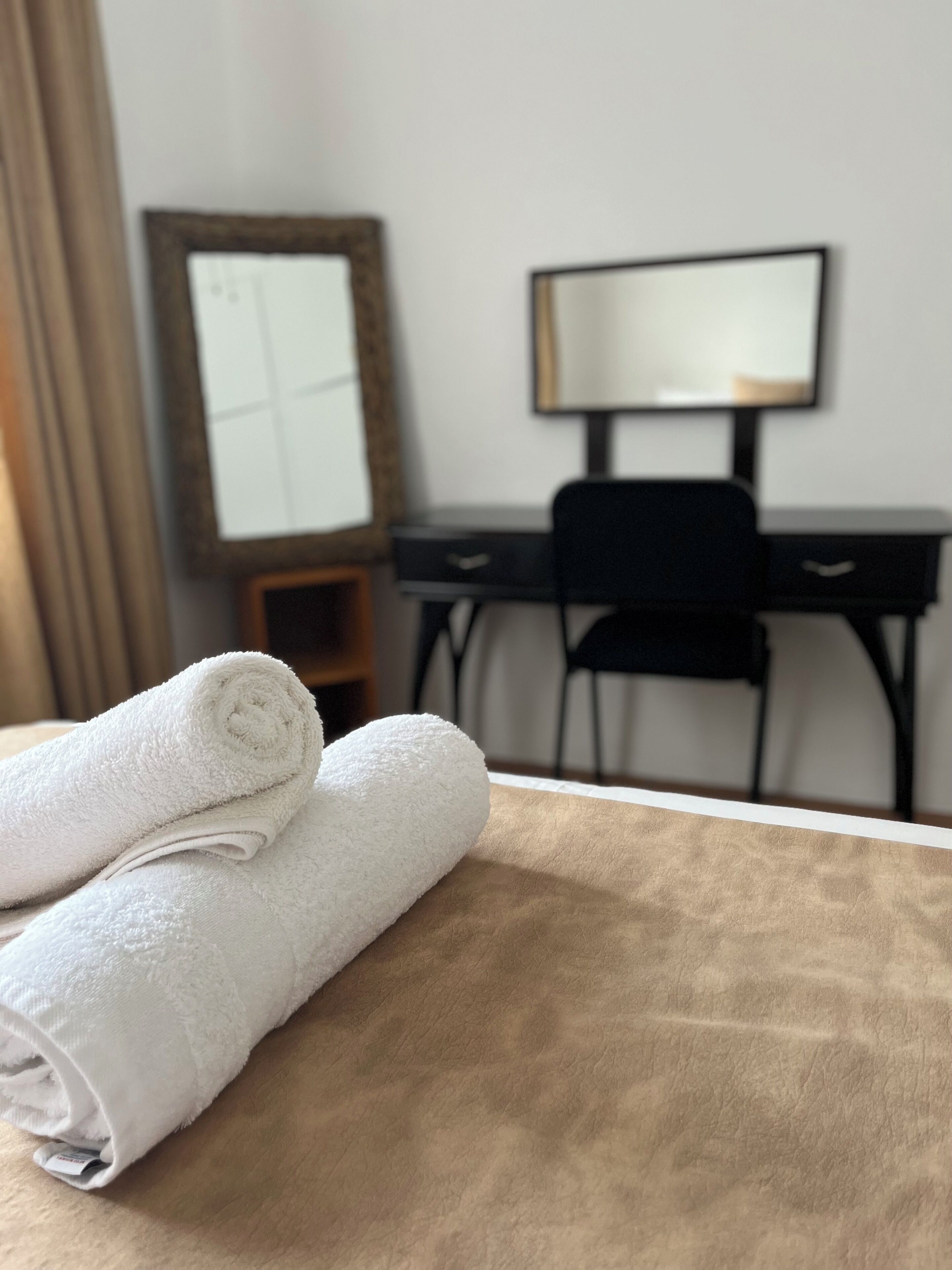 A neatly made bed with white towels, a wooden headboard, a desk with a chair, and a large mirror on the wall.
