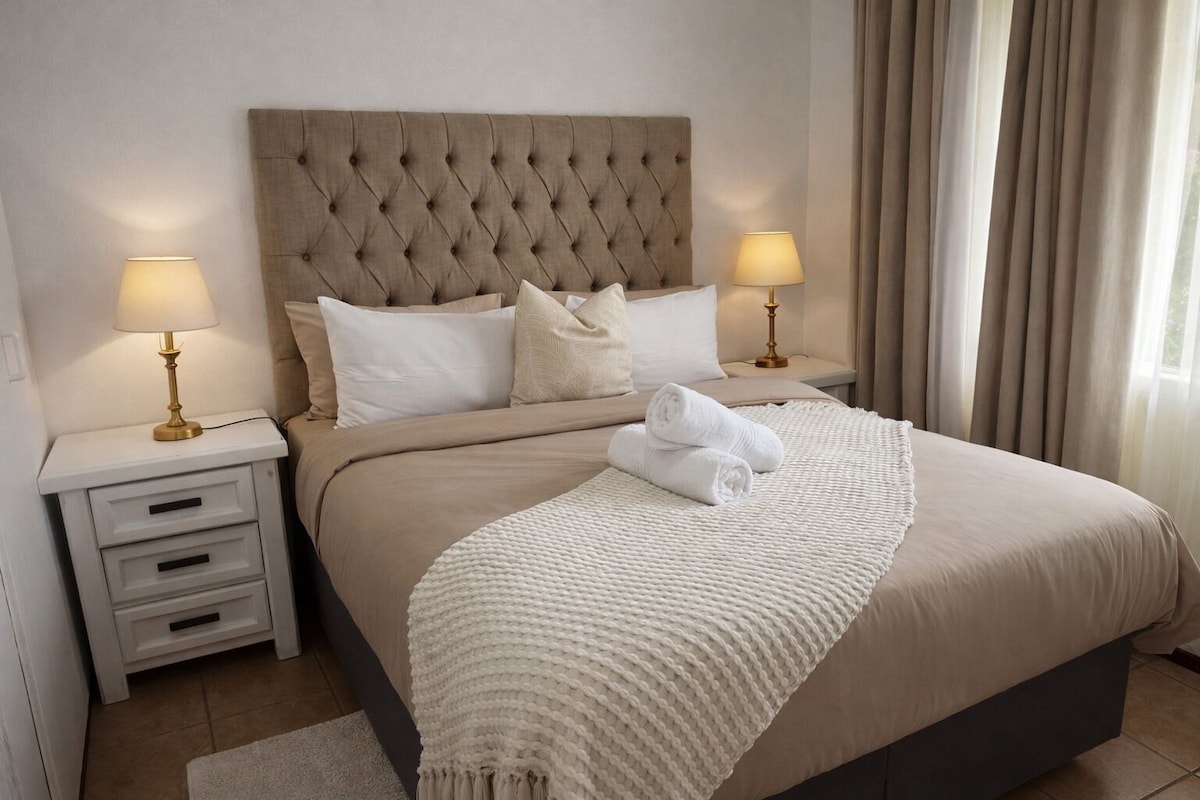 Una cama bien hecha con cabecera capitonada, dos mesitas de noche con lámparas y una ventana con cortinas.