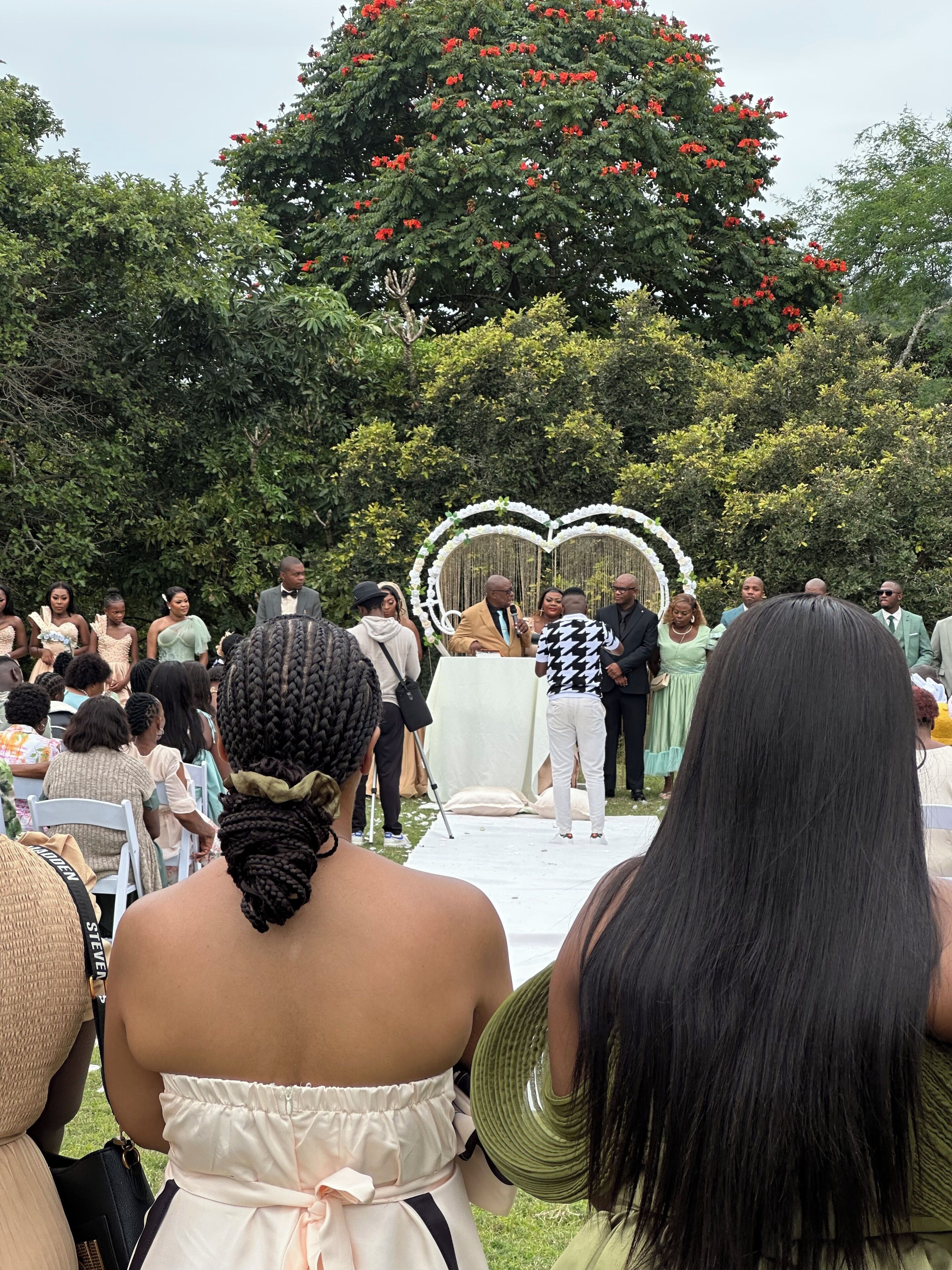 A group of people seated outdoors with a backdrop of trees and a heart-shaped decoration.