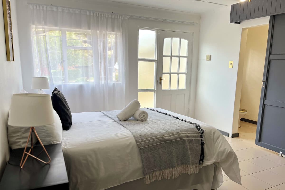 A neatly made bed with a white comforter and a gray blanket, a bedside lamp, and a black pillow on a nightstand.