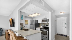 A modern kitchen with white cabinetry, a built-in island, stainless steel appliances, and a flat-screen TV mounted on the wall. - Sandpiper Cove 1016 (Destin)