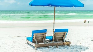 Two wooden lounge chairs with blue cushions and a blue umbrella on a sandy beach with the ocean in the background.