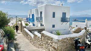 A narrow stone pathway leading to white buildings with blue accents, a parked car, and a clear view of the sea.