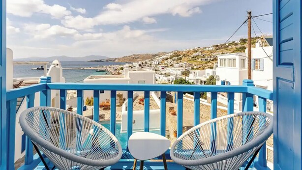 A balcony with two white chairs and a small table, overlooking a coastal town with white buildings and a clear sky.