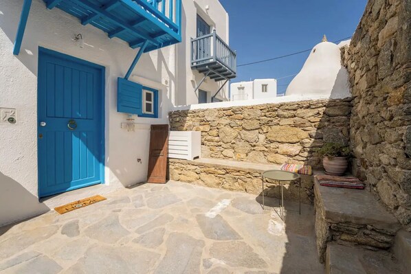 Una calle estrecha con una puerta azul, un muro de piedra y una pequeña mesa con una planta en maceta.