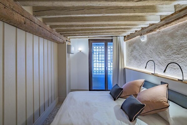 A bedroom with a bed, wooden beams, a window with a view, and a wall-mounted light fixture.