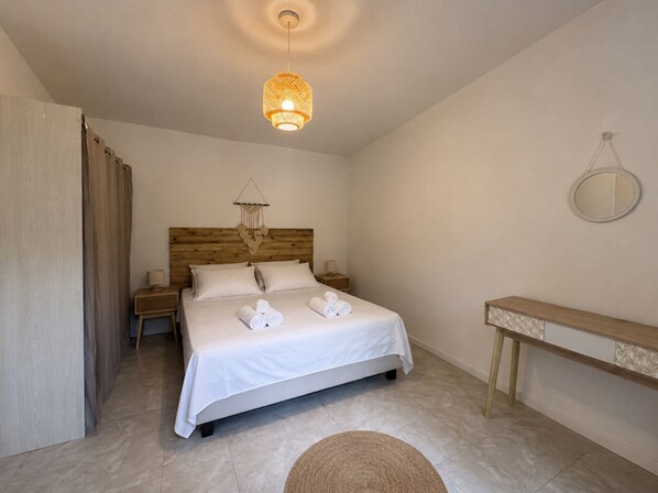 Ein Schlafzimmer mit hölzernem Kopfteil, einem Bett mit weißen Leinen, einem Nachttisch, einer Pendelleuchte und einem kleinen runden Spiegel an der Wand.