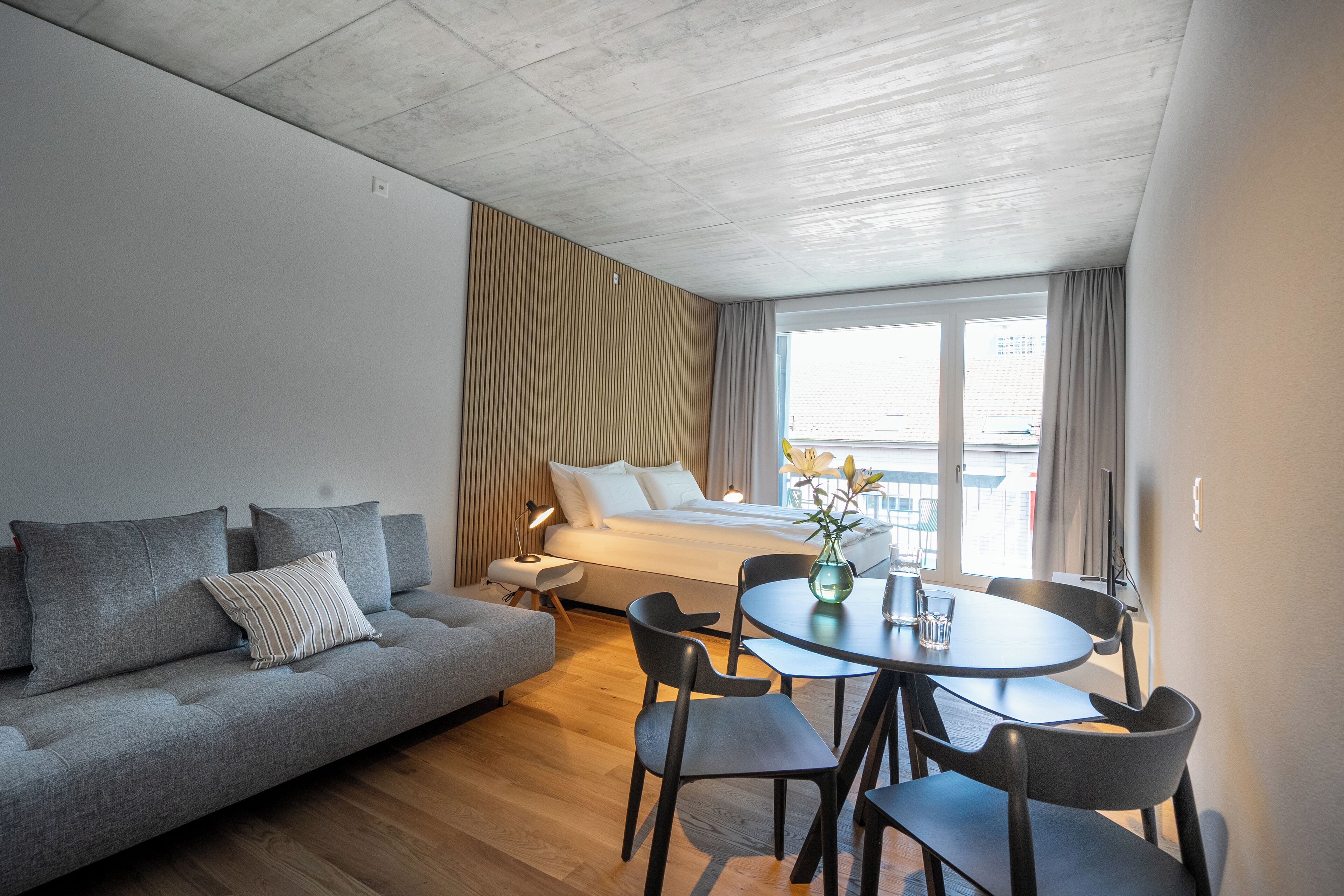 Ein modernes Hotelzimmer mit Bett, grauem Sofa, Esstisch und Stühlen.